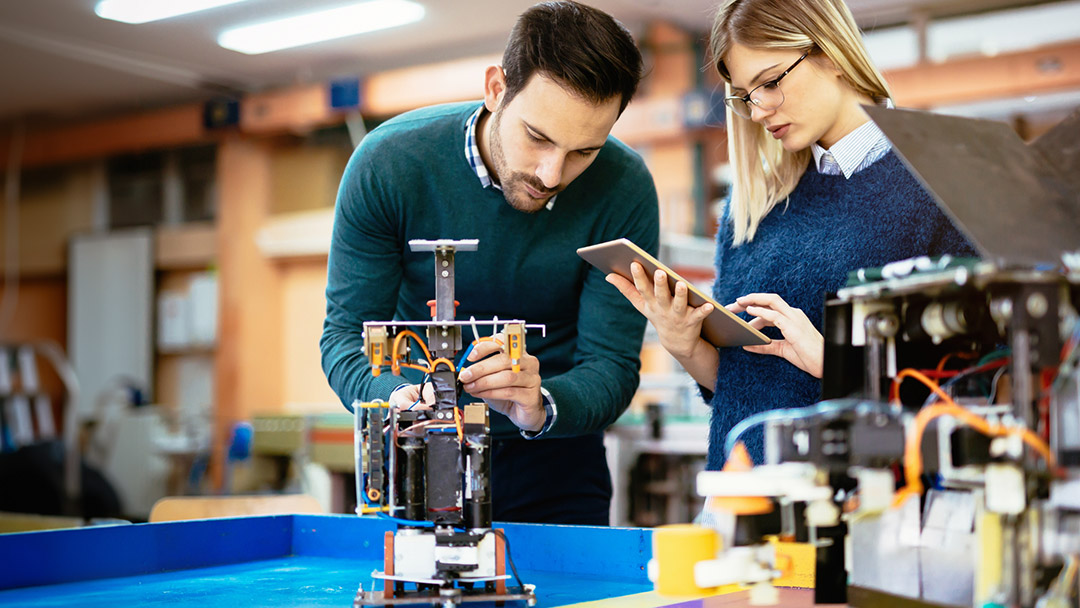 A man and a woman working together in a lab environment, examining a piece of robotic machinery. The woman holds a tablet, while the man adjusts the device, reflecting a collaborative technological setting